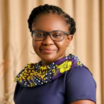 A photo of Patience - she is smiling and wearing glasses, hoop earrings, a dark blue short sleeved shirt and a brightly patterned scarf with yellow, purple navy and white patterns. She has braided black hair that is pulled back.