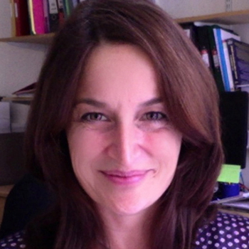 A headshot of Joelle -- she is sitting in an office, smiling and wearing a blue shirt with light polka dots. She has longer light brown hair.