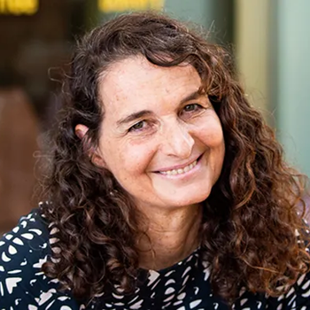 Rachel smiling at the camera in a friendly way. She has long dark brown, curly hair and a black and white blouse.