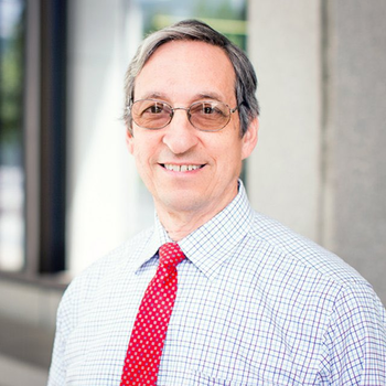 A photo of Thomas. He is wearing a plaid button down (white with blue and burgundy stripes) a red tie, and glasses. He is smiling and has short salt and pepper hair. 