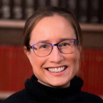 A photo of Catie. She is wearing a black turtleneck and glasses, and she is smiling. She has light brown hair that is pulled back.