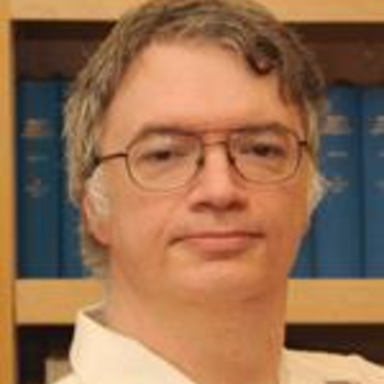 A photo of Travis. He is looking at the camera and lightly smiling. He is wearing glasses and a beige button down collared shirt. He has medium length salt and pepper hair.