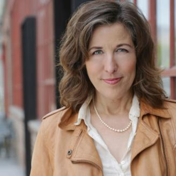 A photo of Laura - she is smiling and wearing a pearl necklace and a white button down shirt with a tan jacket over. She has wavy shoulder length light brown hair.