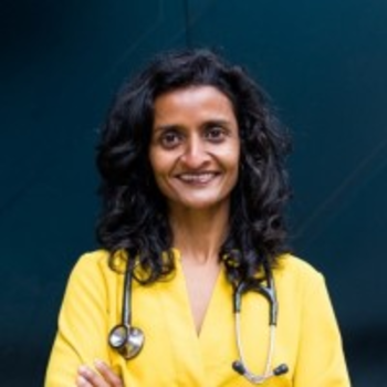 Priya smiling facing the camera directly with her arms crossed. She has curly long black hair and is wearing a yellow shirt. She has a stethoscope around her neck.
