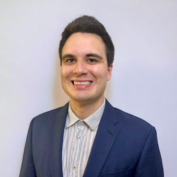 A headshot of Peter - he is wearing a navy suit jacket with a grey button up shirt. He is smiling and has short, dark brown hair
