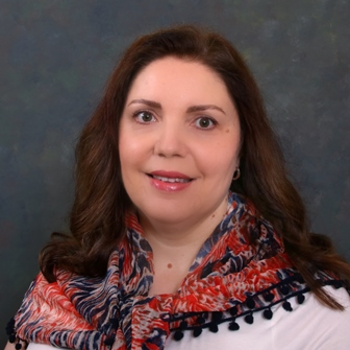 A photo of Lydia. She is looking at the camera and smiling and has long wavy brown hair. She is wearing a white shirt with a colorful blue and red scarf.