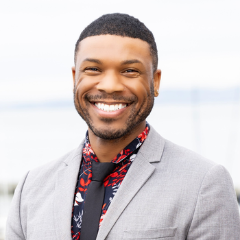 William smiling broadly in to the camera wearing a blue, red, and white patterned shirt with a black tie and grey blazer. He has short cropped dark curly hair.