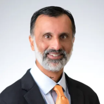 Adithya smiling at the camera. He is wearing a dark suit jacket and orange tie. He has short dark hair and a light grey beard. 