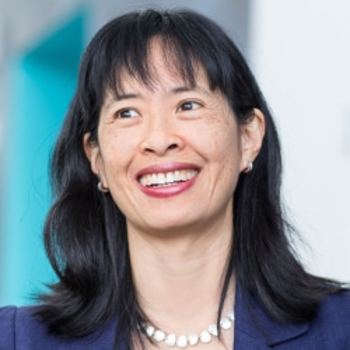 June smiling and looking off camera. She has shoulder length black hair and bangs. She is wearing a dark blue blazer and a white beaded necklace.