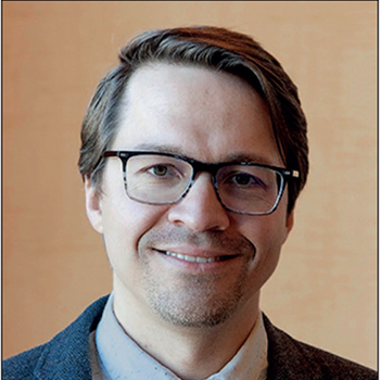 Andrew smiling at the camera. He is wearing glasses and a dark grey suit jacket with maroon tie. He has medium length straight, light brown hair brushed to the right.