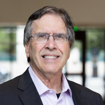 Michael looking at the camera and smiling. He has glasses and short wavy salt and pepper hair. He's wearing a pink button down shirt and a dark grey suit jacket.