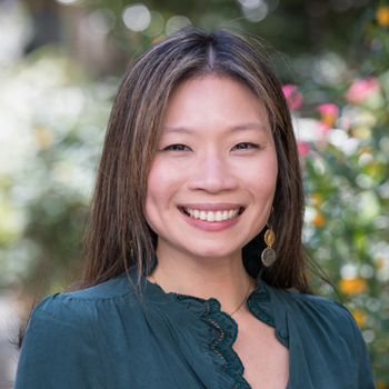Tracy smiling at the camera while standing in front of a flower garden.. She has long light brown hair and is wearing a dark green blouse.
