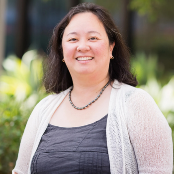 Megie standing outside smiling at the camera. She has long wavy dark hair and is wear a black tank top with a white cardigan over it.