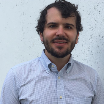 Matthew lightly smiling and looking at the camera. Dressed in a white/blue plaid button down and has a beard and wavy dark hair.